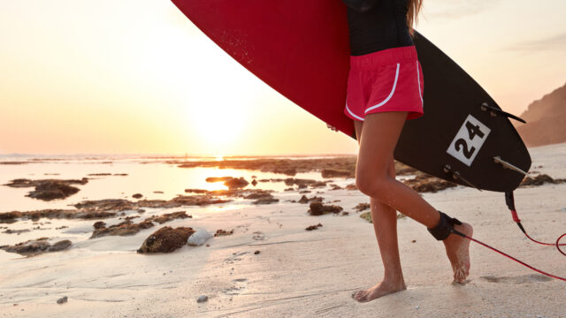 Cropped image of sporty woman in shorts, comes to ocean with surfboard, being fastened with leash, stands on wet sand, beautiful sunset in background, copy space for your advertising content